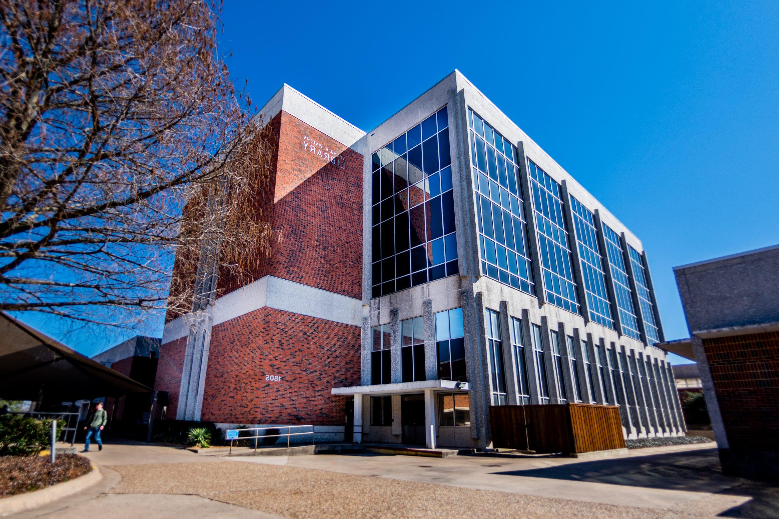 Modern university library building with glass facade and brick exterior. Clear blue sky background. Sign reads "Library" on the side