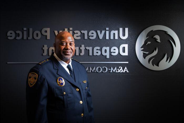 Chief Bryan Vaughn dressed in his dark blue, formal police uniform. He is standing in front of a dark wall with a silver embossed TAMUC lion head logo and the words University Police Department A&M-Commerce.